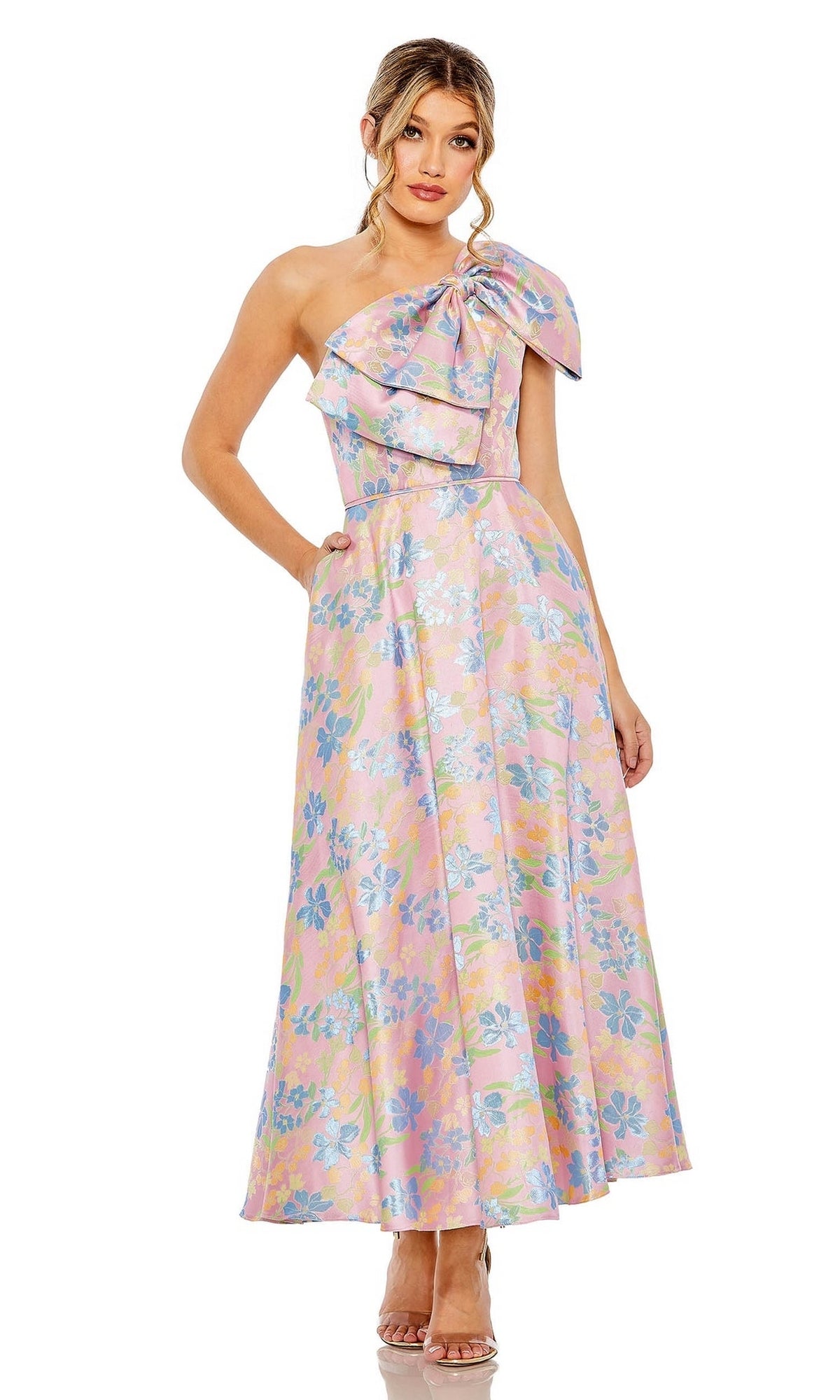 A woman models the Mac Duggal Long Formal Dress 49623, a pink pastel one-shoulder gown with a large bow and ruffle on the bodice, standing with one hand in her pocket and the other at her side.